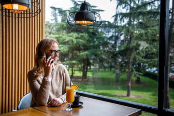 Young charming woman calling with cell telephone while sitting alone in coffee shop during free time, attractive female with cute smile having talking conversation with mobile phone while rest in cafe