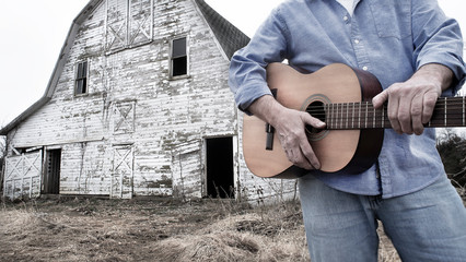 closeup of man holding acoustic guitar standing in front of old abandoned barn on farm in country © Jon