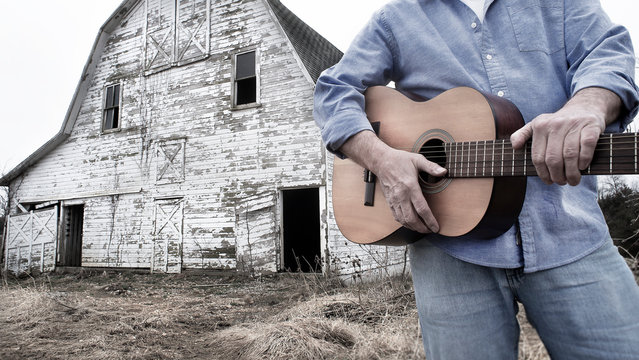 closeup of man holding acoustic guitar standing in front of old abandoned barn on farm in country - Powered by Adobe
