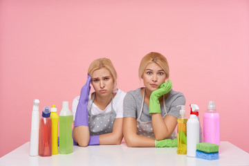 Obraz na płótnie Canvas Indoor photo of pretty young white-headed ladies keeping raised hands on her head while looking at camera with tired faces, sitting over pink background with bottles of household chemicals