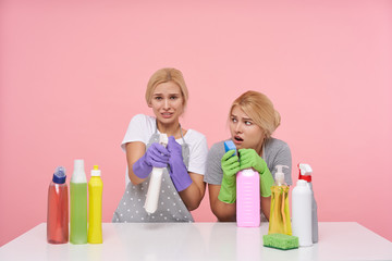Obraz na płótnie Canvas Confused young pretty blonde sisters dressed in working clothes making cleaning and posing over pink background with puzzled faces. Housework and housekeeping concept