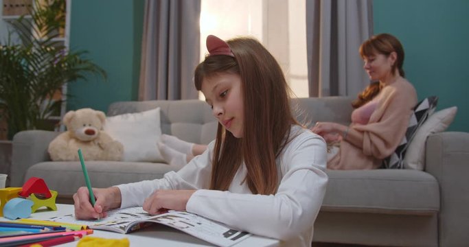 Cute Caucasian girl in funny ears sitting at home and coloring picture while her mother resting on couch and working on laptop computer. Mommy and daughter spending day-off together at home.