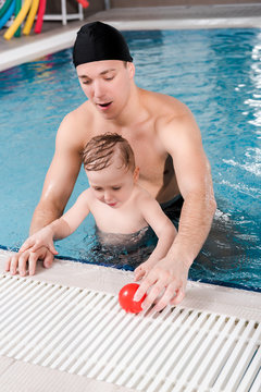 handsome swim coach in swimming cap touching ball near cute toddler boy in swimming pool