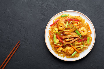 Singapore Mei Fun in white plate on dark slate background. Singapore Noodles is chinese cuisine dish with rice noodles, prawns, char siu pork, carrot, red onion, napa cabbage. Chinese food. Copy space