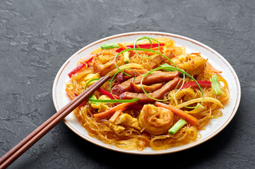 Singapore Mei Fun in white plate on dark slate background. Singapore Noodles is chinese cuisine dish with rice noodles, prawns, char siu pork, carrot, red onion, napa cabbage. Chinese food.