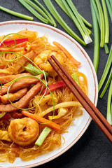 Singapore Mei Fun in white plate on dark slate background. Singapore Noodles is chinese cuisine dish with rice noodles, prawns, char siu pork, carrot, red onion, napa cabbage. Chinese food.