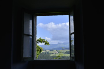 Paisaje verde con bosques y prados y la sierra en el horizonte visto desde el interior de una casa oscura a través de una vieja ventana