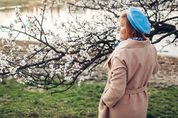 Senior stylish woman walking in spring blooming garden by river admiring nature landscape. Mother's day