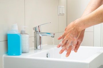 A man washing hands with soap and water in a white square ceramic washstand with bottles of soap and blue alcohol-based hand sanitizer standing beside. Hygiene concept, prevention of viruses’ outbreak