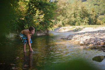 Fototapeta na wymiar A teenage boy in shorts plays in a mountain river in shallow water