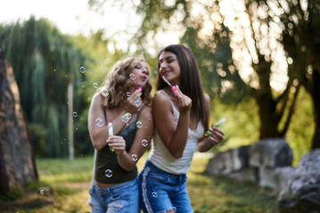 Two sisters, blowing soap bubbles in park, Young pretty girls, wearing jeans shorts, green khaki beige tops, having fun outside. Three-quarter portrait of two girlfriends making soap bubbles, smiling.