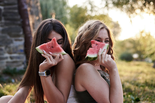 Two sisters, enjoying picnic in city park in summer. Close-up picture of pretty girls,best friends, holding eating watermelon, smiling, laughing. Summertime leisure outside. Active lifestyle.