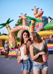 Two sisters, standing is front of attraction in theme park, holding popcorn in paper box, smiling, having fun. Pretty girls. wearing blue jeans and beige and green tops, relaxing in amusement park