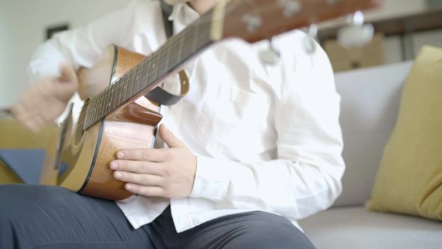 Young guy playing and beating the rhythm on acoustic guitar sitting on cozy sofa near the window. Acoustic guitar practices concept image.