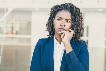 Fototapeta na wymiar Focused pensive leader thinking outside. Serious young black business woman standing at outdoor glass wall, touching chin and looking away into distance. Thinking concept