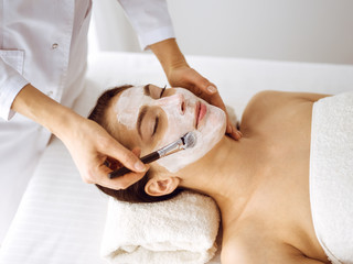 Beautiful brunette woman enjoying applying cosmetic mask with closed eyes. Relaxing treatment in medicine and spa center concepts