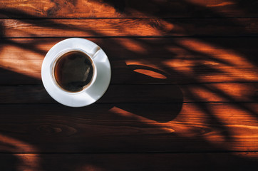 A white cup with coffee and saucer stand on a dark brown wooden mahogany table in the light of the...