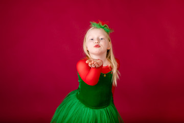 Little blonde girl in a christmas and new year green costume having fun and sending kisses on red background. Space for text. Dreams come true Merry Christmas