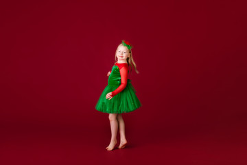 Little blonde girl in a christmas and new year green costume having fun and sending kisses on red background. Space for text. Dreams come true Merry Christmas