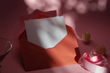 Composition for wedding invitations on a pink background of a terracotta envelope, a white sheet of paper, a pink ribbon, a ring with a stone. Contrasting shadows.
