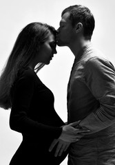 Pregnant girl in black dress and her husband. He hugging her belly, they going to kiss, posing isolated on white. Close up