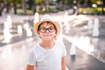 Little caucasian boy in hat and big glasses playing and having fun with water in fountain in the sunny summer park.