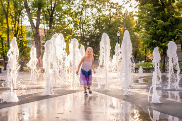 Little caucasian blonde girl in light dress playing and having fun with water in fountain in the sunny summer park.