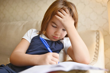 little girl child 7 years old schoolgirl sitting at home on the sofa with a notebook thinking about homework lessons