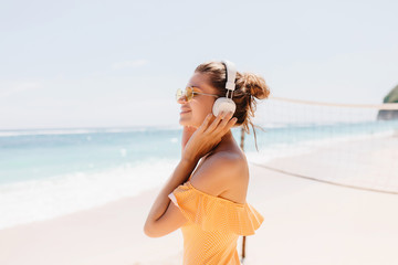 Fototapeta na wymiar Glad smiling woman with tanned skin posing at sea background with charming smile. Outdoor portrait of enthusiastic girl wears big white headphones while chilling at ocean coast.