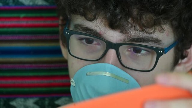 Portrait of a bedridden caucasian boy wearing a blue protective mask. He is studying his orange-covered notebook. Home study concept during coronavirus pandemic quarantine.