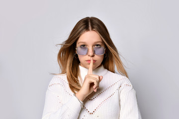 Blonde adolescent in sunglasses, smart watch, bracelet and sweater. She showing be quiet sign, posing isolated on white. Close up