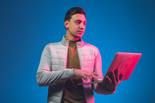 Using Laptop. Caucasian Man's Portrait Isolated On Blue Studio Background In Pink Neon Light. Beautiful Male Model In Casual. Concept Of Human Emotions, Facial Expression, Sales, Ad. Copyspace.