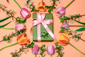 Round frame made of colorful pink and orange tulips with cherry blossom twigs on peachy background and gift box wrapping in green paper with pink ribbon. Beautiful spring floral greeting card