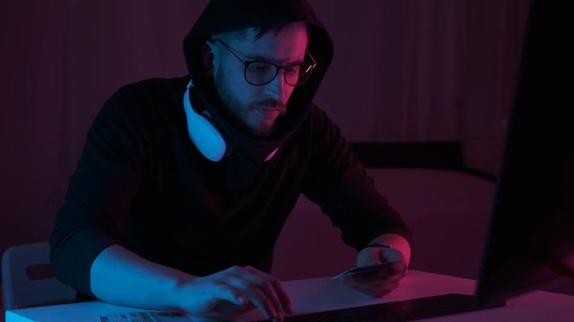 Cyber crimes and cybercriminals. Dangerous hacker breaks into banking cards. Man in a hood hacking secret program or website systems in dark room. Cyber attack. Programming on screen laptop. closeup