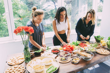 Young women preparing healthy meal. High angle view of beautiful young women in sportswear standing near table and cooking fresh organic vegetables. Healthy eating concept