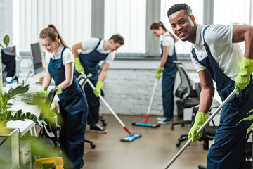 selective focus of smiling multicultural team of cleaners washing floor with mops in office - obrazy, fototapety, plakaty