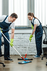 two young cheerful cleaners in uniform washing floor with mops in office