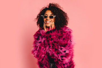 Happy young african  woman excited touching face in pink faux fur coat at pink background in...