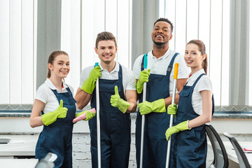 happy multicultural team of cleaners looking at camera and showing thumbs up