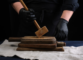 male cook in black uniform holds a wooden hammer for beating meat on a vintage cutting board