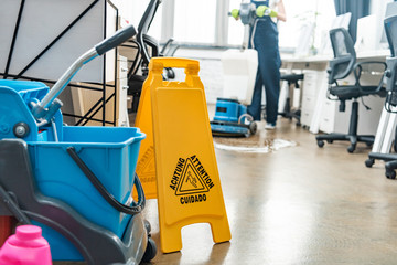 selective focus of cart with buckets and wet floor caution sign, and cleaner washing floor with cleaning machine on background