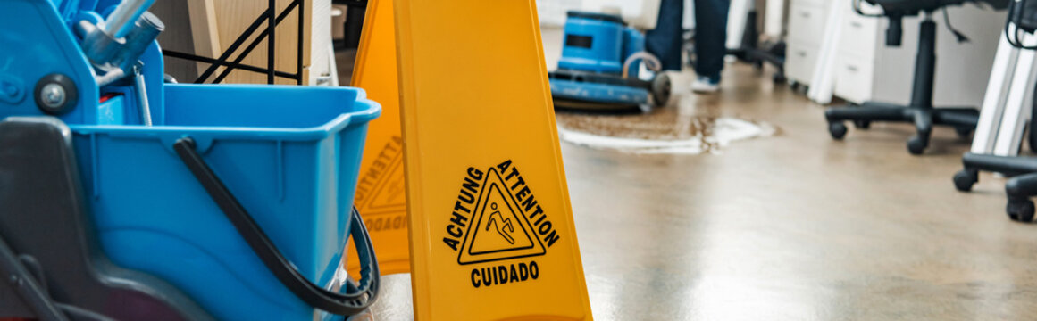 selective focus of wet floor caution sign and bucket, while cleaner washing floor with cleaning machine on backround
