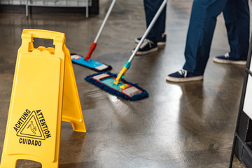 partial view of two cleaners washing floor with mops near wet floor caution sign