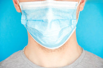 A man in a blue medical mask looks at the camera, sideways. A man in a gray T-shirt on a blue background. Place for text.  Copy Space. Covid-19