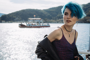 Portrait of a girl with blue hair in a purple dress and a black jacket on a background of a bay by the mountains
