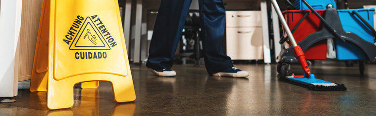 cropped view of cleaner washing floor with mop near wet floor caution sign, panoramic shot
