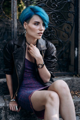Fototapeta premium Portrait of a girl in a dress and a black leather jacket sitting on the steps near the forged gate