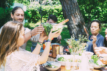 Cheerful young people at table in yard. Happy young male and female friends gathering around table with tasty meal and drinks in backyard. Healthy meal concept