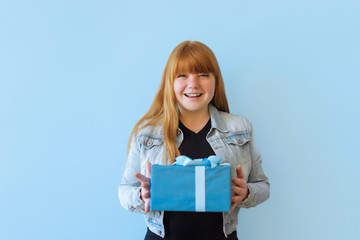 Obraz premium a happy woman received a blue gift box with a bow. She is happy and flattered by attention. Girl dancing on a blue background. she is wearing a denim jacket