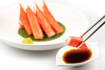 Stainless steel chopsticks are squeezing Crab Stick Sashimi in a white dish with dipping sauce with tamari shoyu and wasabi.